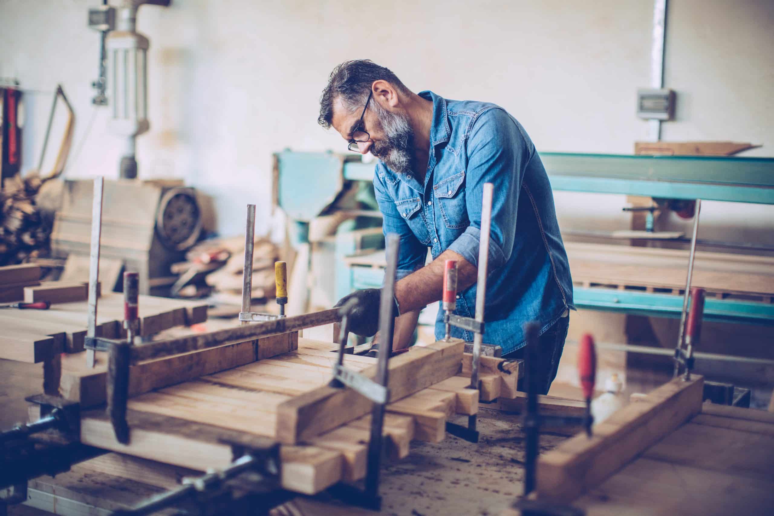 A craftsman making wooden table on workbench. He's using carpenter press to fix wooden planks. Workshop in background, lot of tools and crafts of different type.
