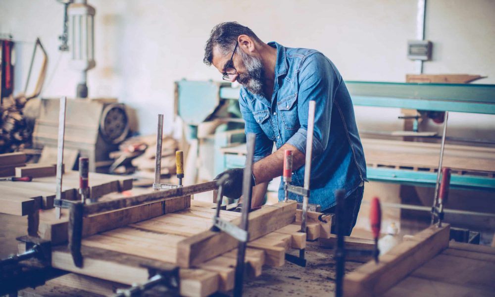 Master woodworker A craftsman making wooden table on workbench. He's using carpenter press to fix wooden planks. Workshop in background, lot of tools and crafts of different type.